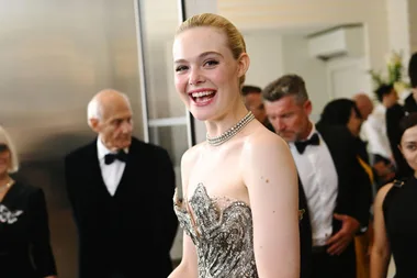 A person smiles in a sparkly strapless gown with a pearl necklace at Cannes 2023 event, surrounded by people in formal wear.