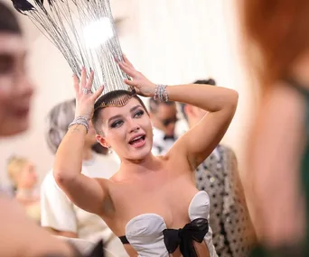 A woman in a strapless gown with a dramatic headpiece adjusts it, surrounded by people at a formal event.