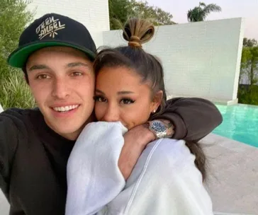 A couple hugging by a pool, smiling; woman hides part of her face with a sleeve, man wears a cap.