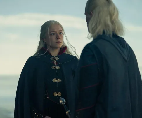 Two characters with long blonde hair in medieval attire stand facing each other against a cloudy sky backdrop.