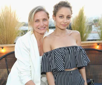 Two women smiling at an outdoor event, one in a white suit and the other in a black and white striped outfit.