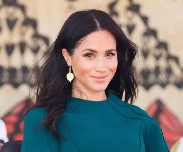 A woman with long dark hair, wearing a teal top and leaf-shaped earrings, smiling outdoors.