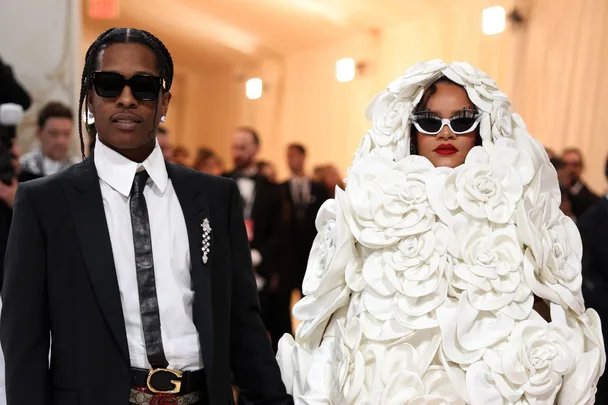 Man in black suit and woman in a floral white gown with sunglasses at Met Gala 2023.