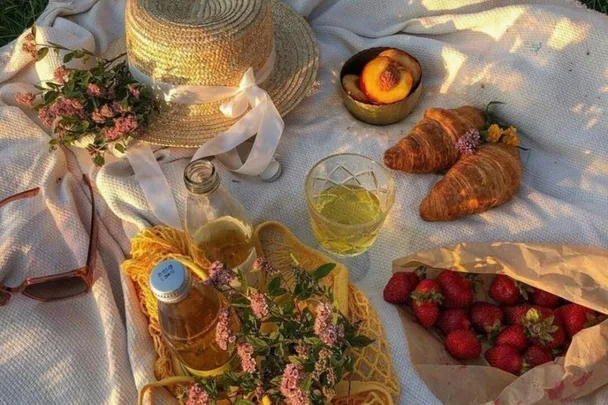 Picnic setup with a hat, flowers, croissants, peaches, strawberries, juice bottle, glass, and sunglasses on a blanket.