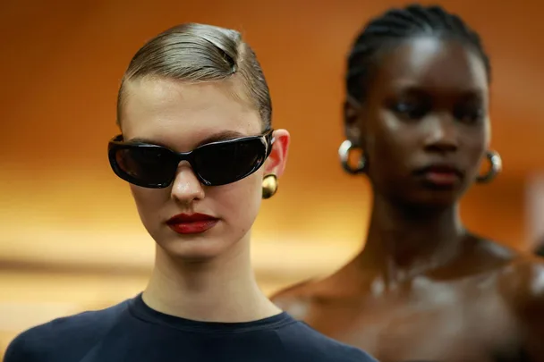 Women in sunglasses and bold makeup, wearing gold accessories, with an orange background.