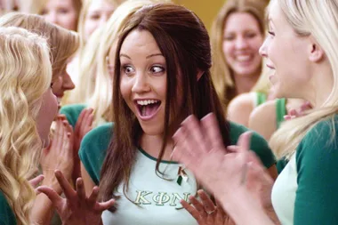 Excited woman in a sorority setting, surrounded by cheering people, wearing a shirt with Greek letters.