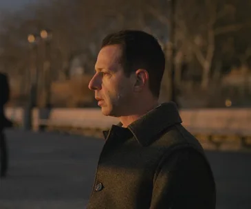 A man in profile stands outdoors at sunset, wearing a dark coat, with blurred trees in the background.