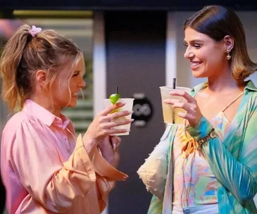 Two women in colorful outfits smiling and clinking their drinks together.