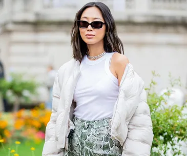 A person wearing sunglasses, a white puffer jacket, a sleeveless top, and patterned pants stands outdoors.