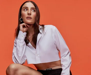 Woman with long hair in a cropped white shirt poses against an orange background.