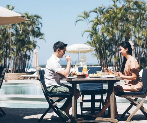 Two people dining outdoors near a pool with tropical plants in the background, enjoying a meal under a clear sky.