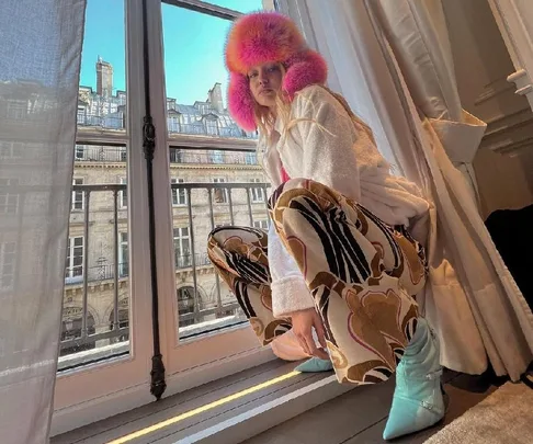 Person wearing a vibrant pink hat and patterned pants, crouching by a window with a cityscape view outside.