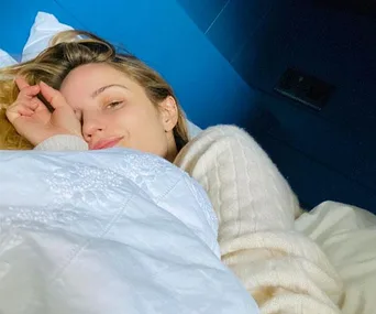 Person smiling while lying in bed under a white blanket in a blue room.