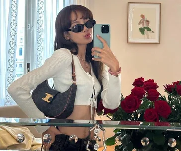 Person taking a mirror selfie, wearing sunglasses and a white top, holding an Ava bag, with a bouquet of red roses nearby.