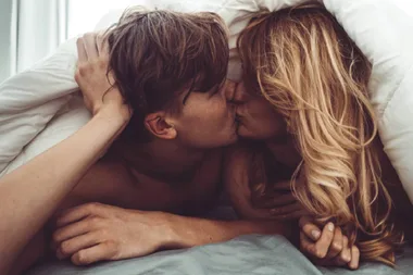 A couple kissing under a duvet, lying closely on a bed, with soft light filtering through a window.