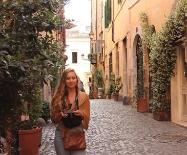 Woman holding a camera in a quaint, cobblestone European street lined with plants and charming buildings.