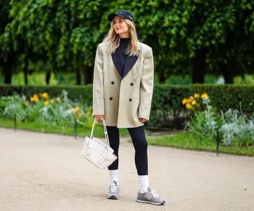 Woman in oversized blazer, black leggings, cap, and sneakers holds a white bag, standing in a park with greenery.