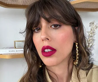 Woman with wavy brown hair and fringes, bold pink lipstick, wearing large gold hoop earrings.