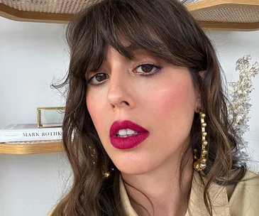 Woman with wavy brown hair and fringes, bold pink lipstick, wearing large gold hoop earrings.