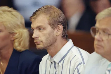 A man in a striped shirt sits between two others in a courtroom setting, focused and looking forward.