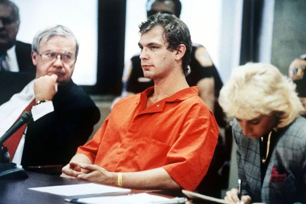 A man in a prison jumpsuit sits attentively in court between two people, one writing notes.