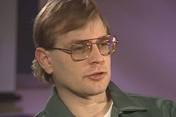 A person with glasses and short hair wearing a green jacket, appearing to be in a contemplative pose.