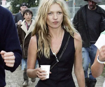 A woman with long blonde hair wearing a black vest holds a cup while walking outdoors at a festival.