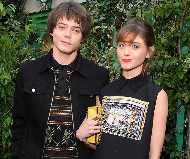 A man and woman posing together outdoors, both wearing stylish clothing, with greenery in the background.