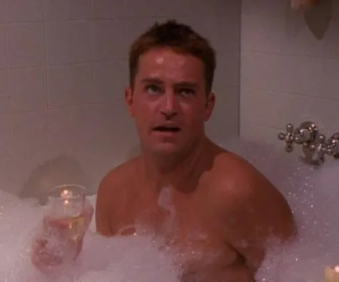 Person in a bubble bath holding a drink, looking upward, surrounded by tiled walls.