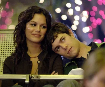 A woman and a man sitting closely, the man rests his head on the woman's shoulder, with colorful lights in the background.