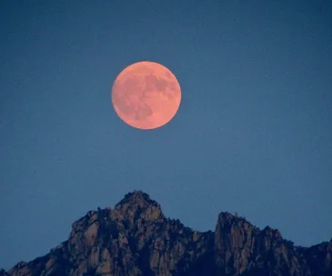 Reddish full moon rising over jagged mountain peaks against a deep blue sky.