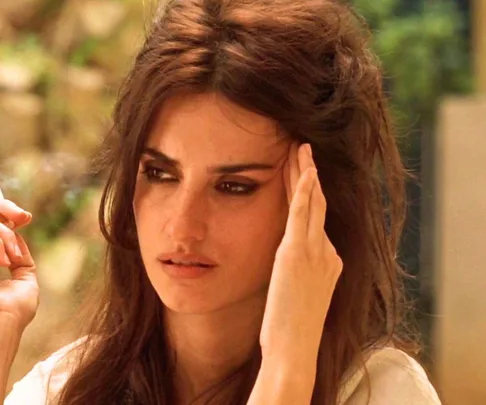 A woman with long dark hair and dramatic eye makeup, holding her head in thought against a blurred outdoor backdrop.