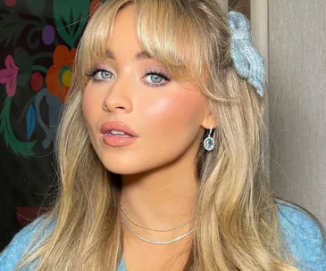 Portrait of a woman with long blonde hair, wearing blue hair bow and earrings, with vibrant floral background.