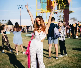 Festival-goer in a white jumpsuit and sunglasses, arms raised, at an outdoor event with colorful structures and crowds.