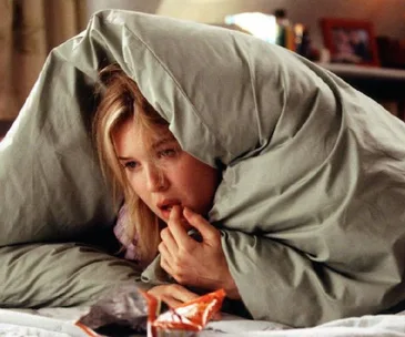 Woman in bed eating snacks under a blanket, looking pensive in a cozy bedroom setting.