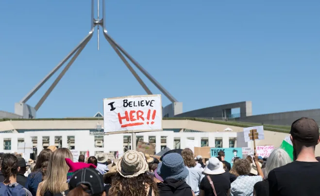 Protesters outside a government building, one holding a sign reading "I BELIEVE HER!!"