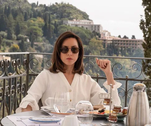 Person in white blouse and sunglasses sits at an outdoor table with breakfast, overlooking a lush, hilly landscape.