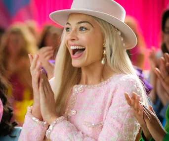 Barbie in a pink outfit with matching hat, clapping and smiling joyfully.