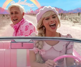 Barbie and Ken smile while driving a pink convertible through a vibrant, desert landscape with a rainbow overhead.