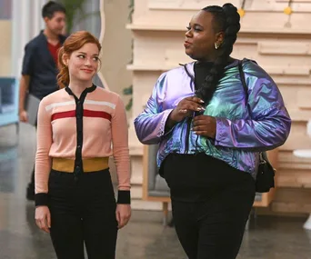 Two people walking indoors, one in a pink sweater and the other in a shiny purple jacket, engaged in conversation.