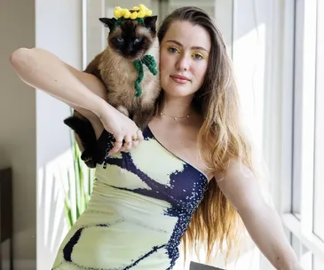 Person in a colorful dress with a Siamese cat on their shoulder near a bright window.