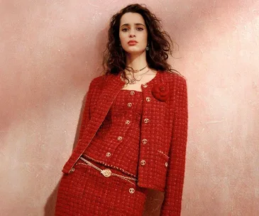 Model in red tweed suit with gold chains, featuring a floral brooch, against a textured pink background. Chanel 2023-2024.