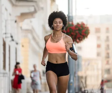 A woman jogging outdoors, wearing a peach sports bra and black shorts, listening to music with earbuds.