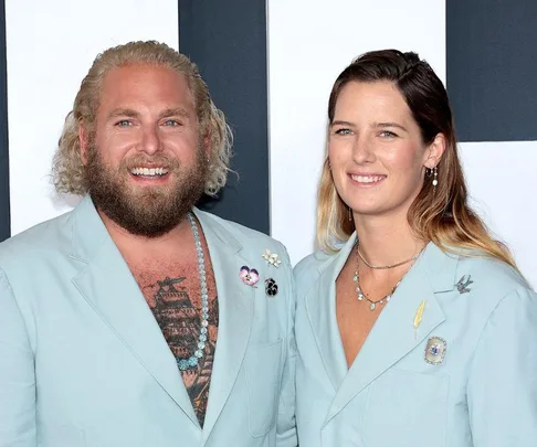 A man and woman smiling in matching light blue outfits with pin accessories on a striped background.