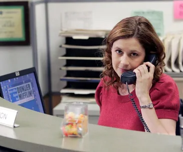 A woman with curly hair, wearing a red top, sits at an office desk holding a phone, by a "Dunder Mifflin" computer screen.