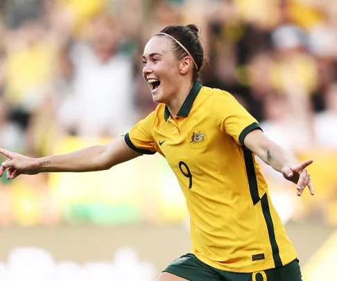 Soccer player in yellow Matildas jersey celebrates with arms outstretched during a match.