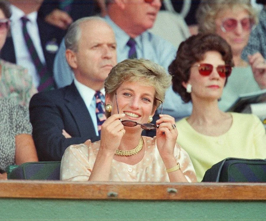 A woman in a light outfit smiles, holding sunglasses, surrounded by spectators at a tennis event.