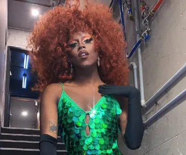 Drag queen in a vibrant green sequin dress and large curly red hair, posing in a backstage hallway.