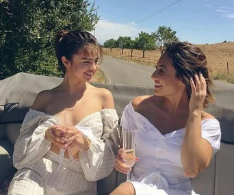 Two women in off-shoulder dresses enjoy a conversation and drinks on a sunny day.