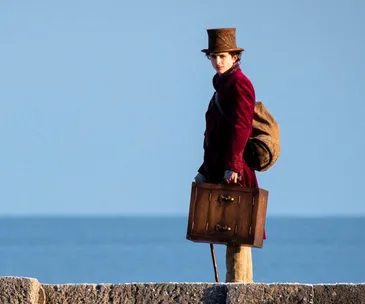 Young Willy Wonka stands on stone pier with suitcase, wearing red coat and top hat, against blue sky and sea background.